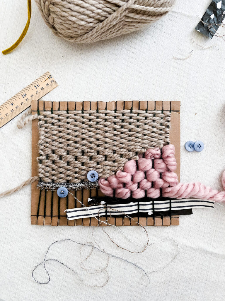 A photograph of a partially completed weaving consisting of multiple types of material including brown yarn, thick pink yarn, and black and white ribbon. 